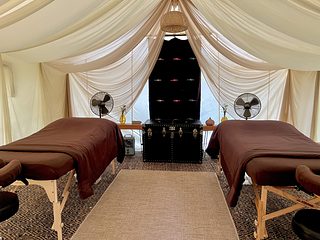 A spacious canvas tent set up as a massage room, with two massage tables covered in blankets, small side benches, a patterned rug, and soft daylight filtering through the tent walls.
